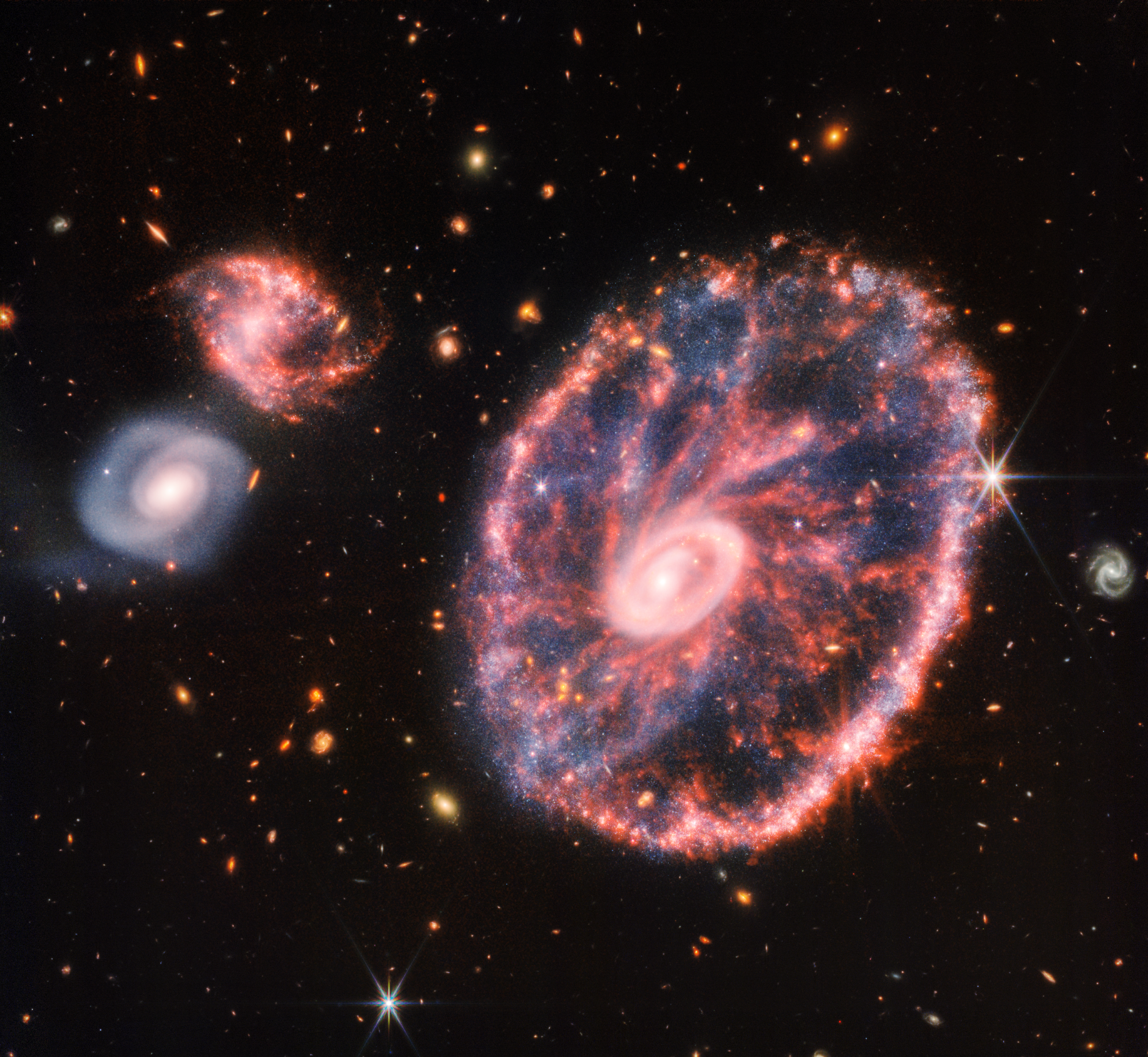 A large pink, speckled galaxy resembling a wheel with a small, inner oval, with dusty blue in between on the right, with two smaller spiral galaxies about the same size to the left against a black background.